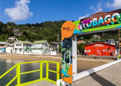 Colorful welcome to Tobago sign at the Man of War Bay pier with The Cholson Chalets eco-luxury beachfront property in the background